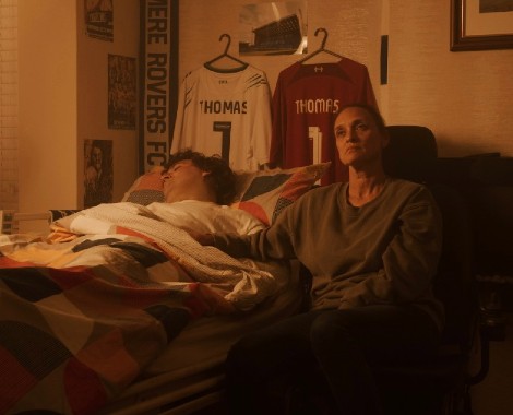 A woman sits beside a man, who is lying sick in bed. Two football shirts with 'Thomas 1' printed on the back hang on the wall behind.
